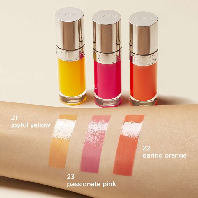 Presentation of the Clarins lip oil range in dark orange, dark orange, and orange, with the colors swatched on a woman's arm